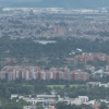 Vista panorámica de Bogotá bajo cielo nublado. En primer plano se observan edificios residenciales rodeados de árboles y zonas verdes, mientras al fondo se aprecia una amplia extensión urbana con edificaciones de diferentes alturas cubiertas por una ligera neblina.