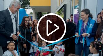 Momento de inauguración. Dos adultos y tres niños pequeños sostienen una cinta para dar apertura a un jardín infantil.